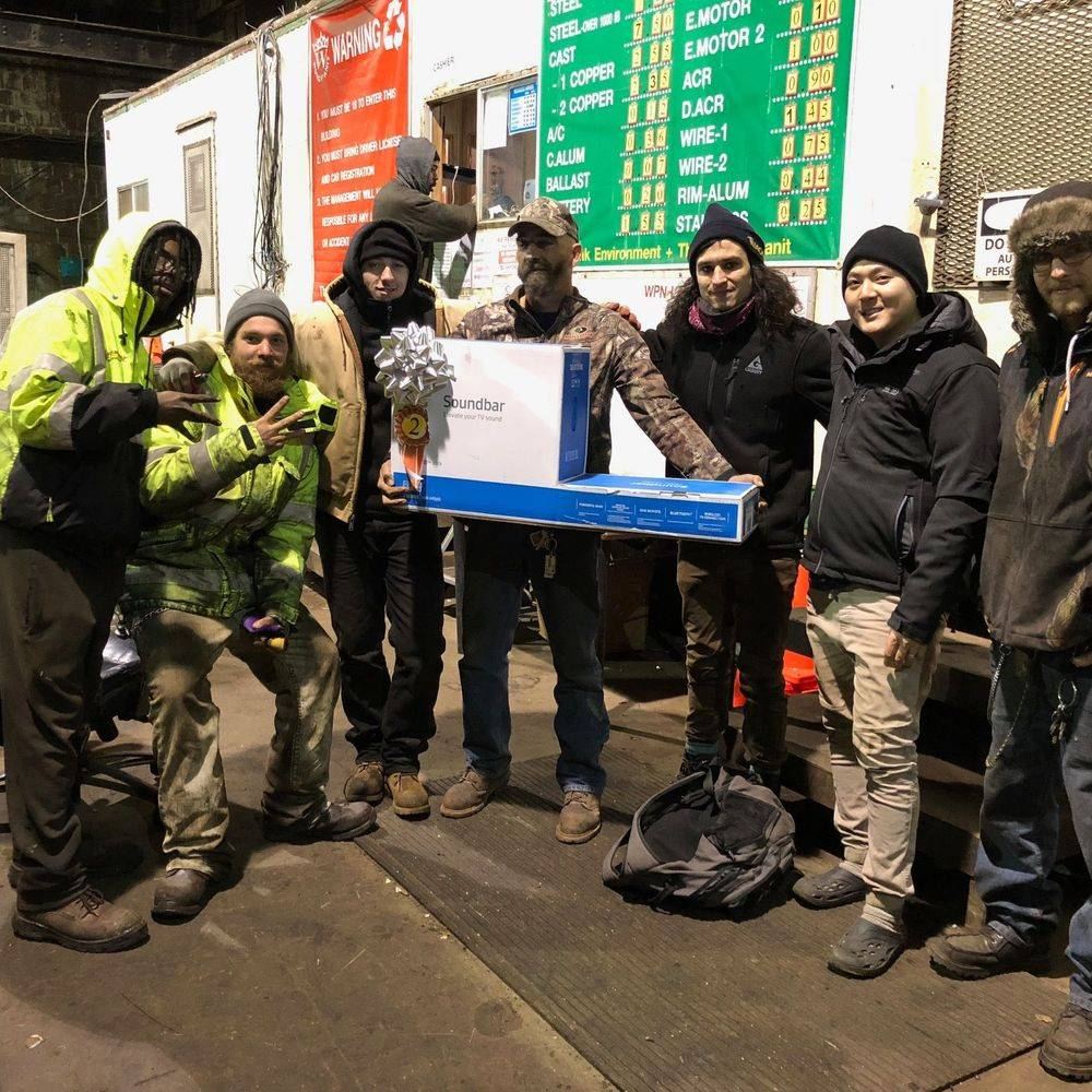 Seven people standing in the warehouse holding a boxed television, in front of the price board.